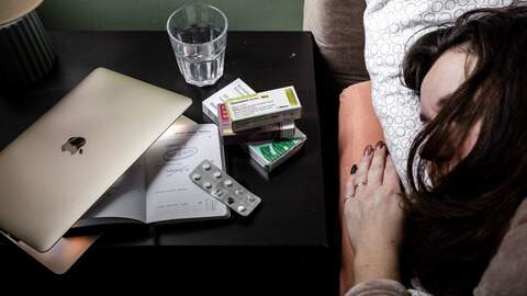 Een vrouw ligt in bed met een dichtgeklapte laptop, agenda met geschrapte werkdagen en antidepressiva medicijnen op het nachtkastje. 