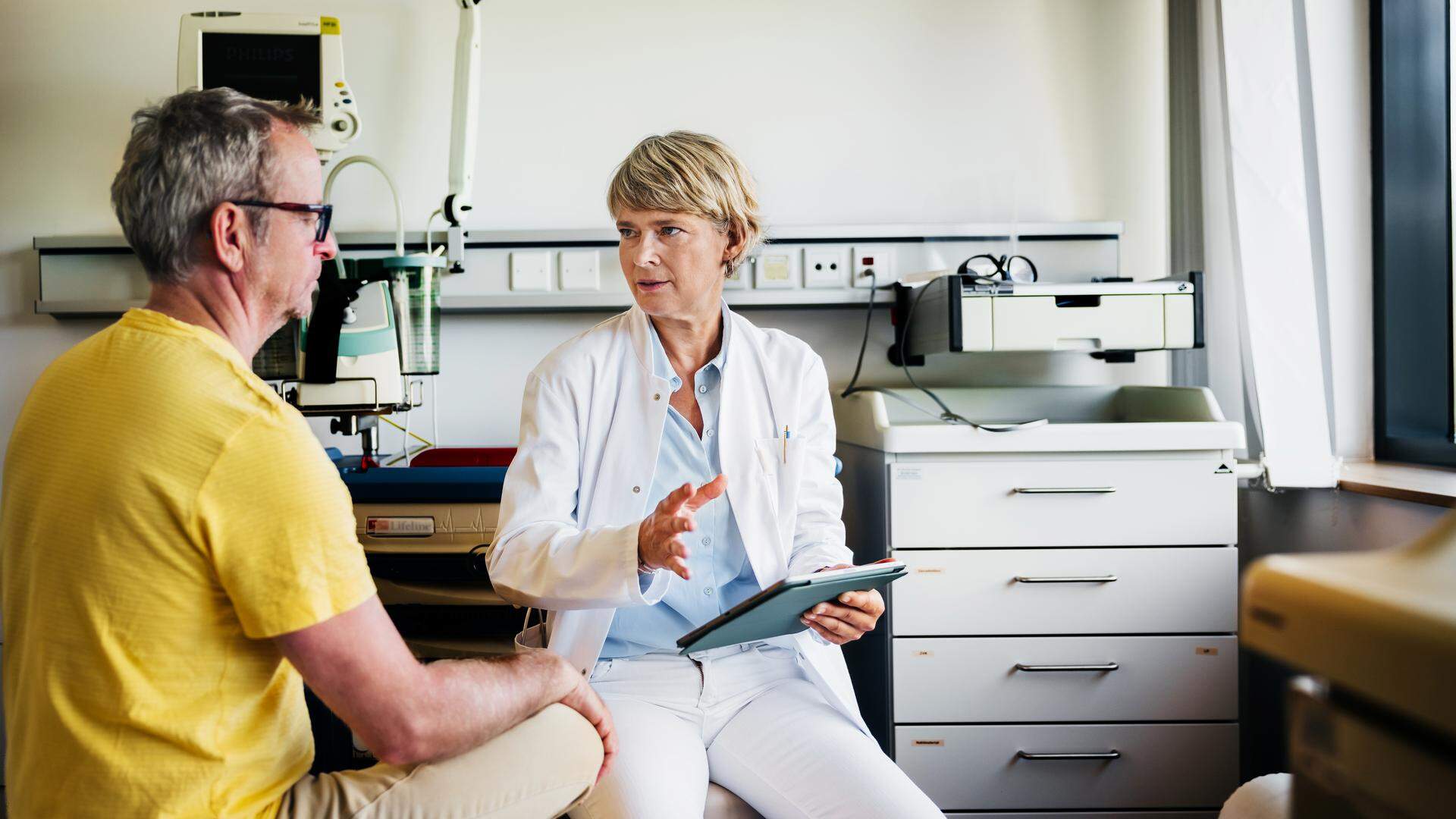 A female doctor in a clinical setting shows a digital X-ray to a patient while engaging in a discussion. The patient, dressed in a yellow shirt, listens attentively, seated across from the doctor.